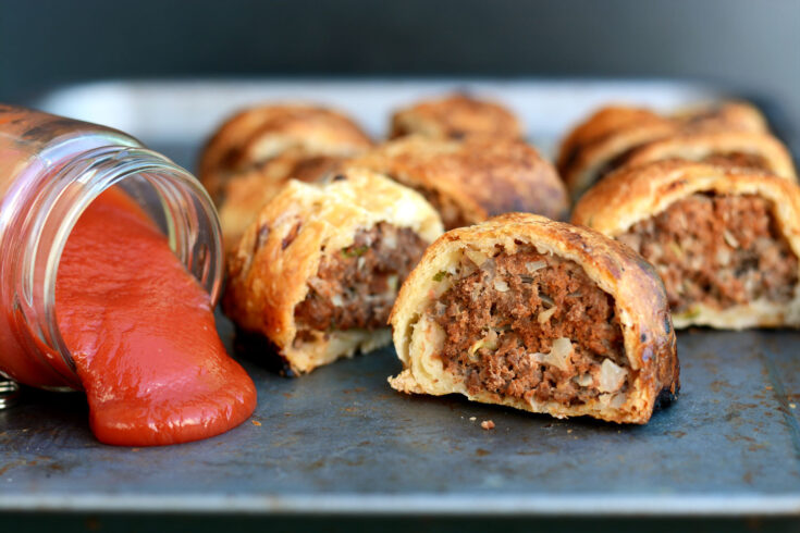 Australian sausage roll on a metal plate with a jar of ketchup (tomato sauce) knocked over next to it.
