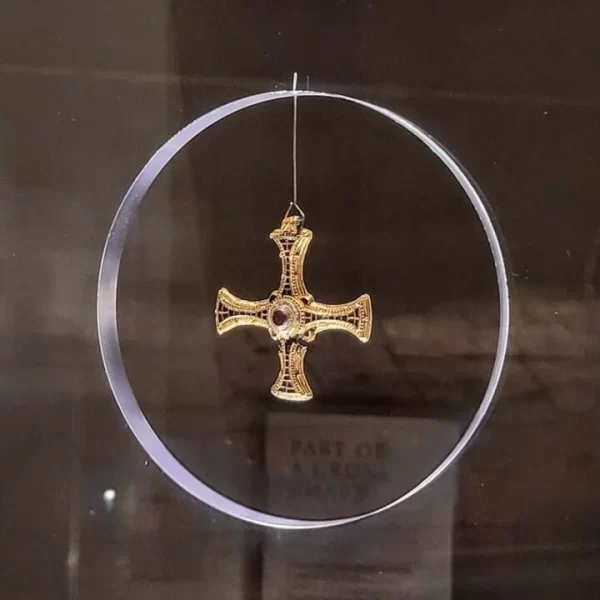 Close up image of gold cross with red jewel in centre. The cross is displayed hanging in the centre of a circular frame