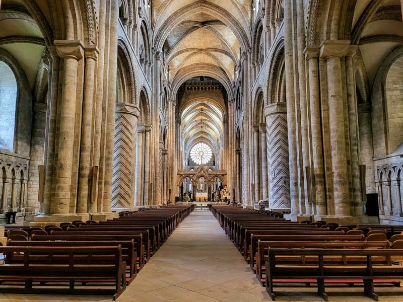 Wide view of interior of Durham Cathedral. Soaring pillars on the left and right sides and circular stained-glass window in the middle rear of the photo. Just one of many things to do in Durham for couples