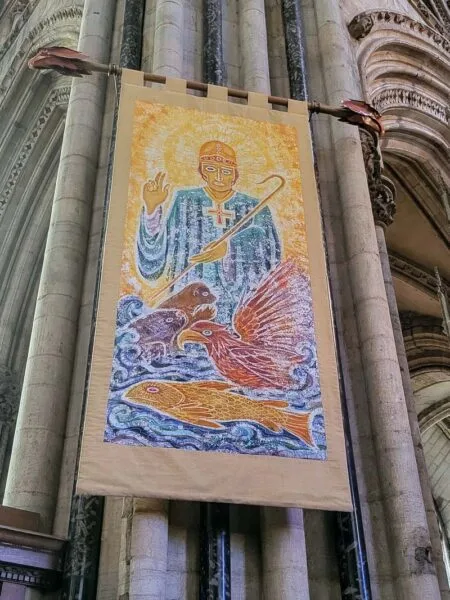 Banner set on interior pillar. Yellow background. St Cuthbert dressed in blue with animals in lower half of banner