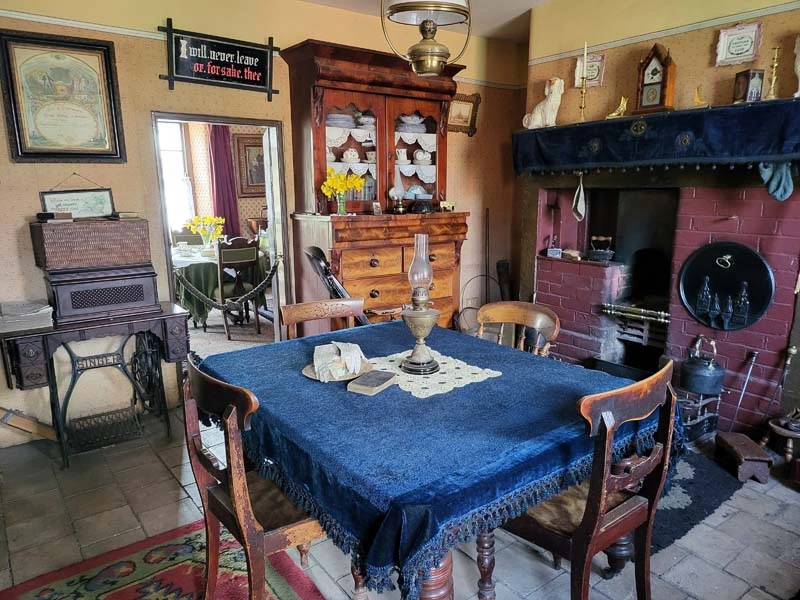 Interior of miners home. Kitchen. Dining room table with blue tablecloth, wooden cabinet with china in centre rear of image. 