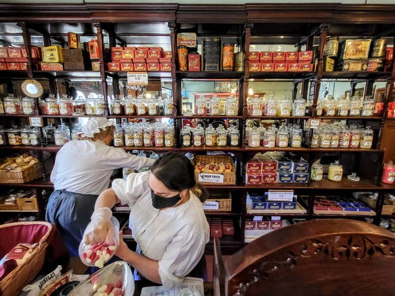 Interior of Jubilee Confectioners. Large mirrored shelves run left to run full of jars of sweets. Two shop assistants can be seen lower left. One reaching for a jar on the shelf and one pouring sweets into a weighing scale. 