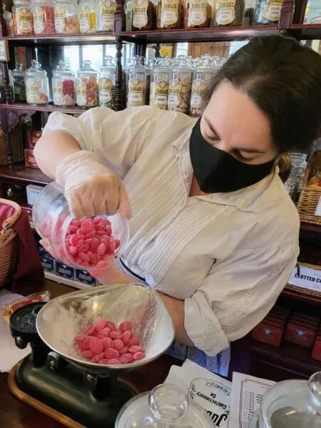 Close up of shop assistant pouring sweets from a glass jar into a traditional weighing scale with weights. 