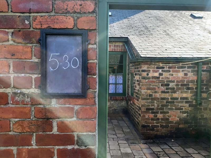 Exterior photo of front entrance of miner's house. Brick wall fills half the left side of photo. On the brick wall is a small rectangular chalk board with the numbers "5:30" written on it. The stone courtyard and house fill the right side of photo