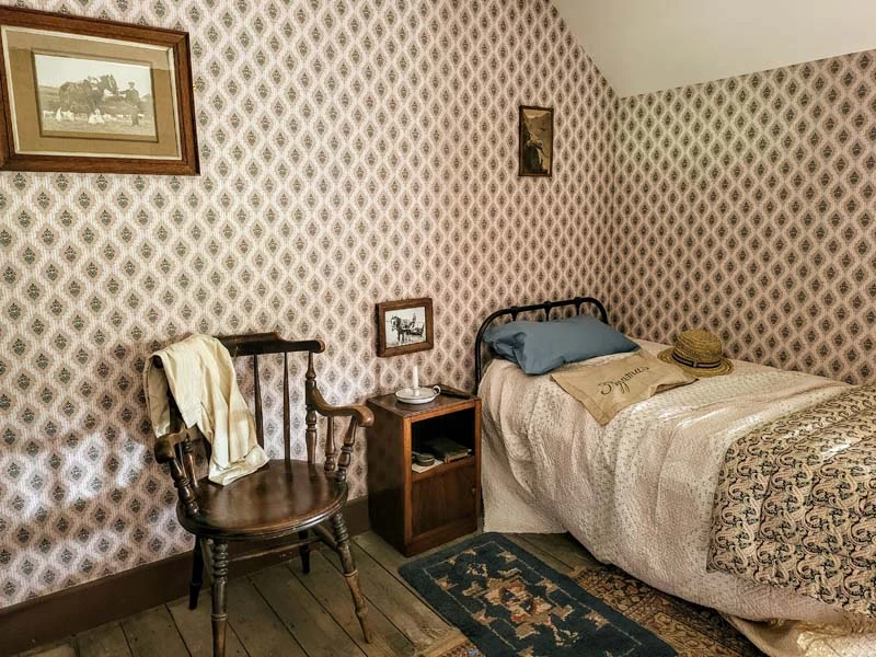 Interior of farm house bedroom. Bed to the right of the bedroom and brown wooden chair to the left centre. Picture frame hangs on wall in the top left of image. 