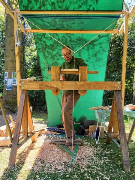 Photo of Chris standing behind the shave horse as he crafts his design. A green tarpaulin can be seen covering Chris and the workspace, supported by a timber frame.  