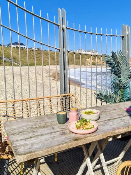 A wooden table in middle of photo, with brunch items set atop it, and against a background of an iron fence and seaside views