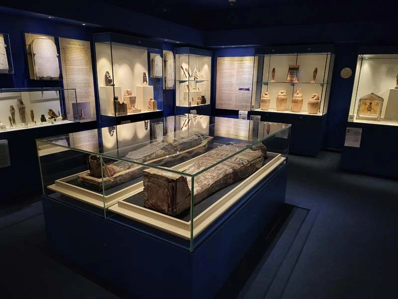 Interior of Ancient Egypt Gallery. Wide-angle photo with sarcophagus in the centre of photo and surrounded by display cases containing artefacts in the background. 