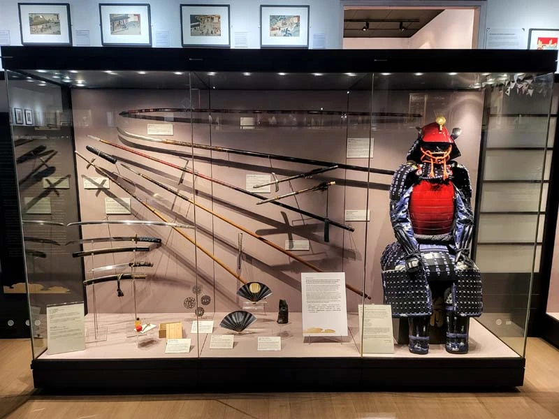 Interior of museum. Large glass display case. The right side of photo ois the full suit of Samurai armour, red and blue. The left side of display case are various swords and other weapons.