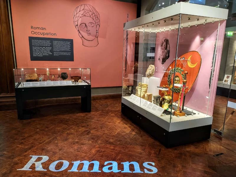 Roman artefacts in glass cabinets to the right side and the word "Romans" on the bottom of image"