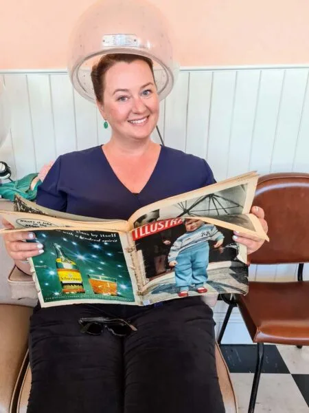 Photo of Roma sitting in 1950s hairdressing chair under a traditional hairdryer. Roma is smiling whilst reading a 1950s magazine