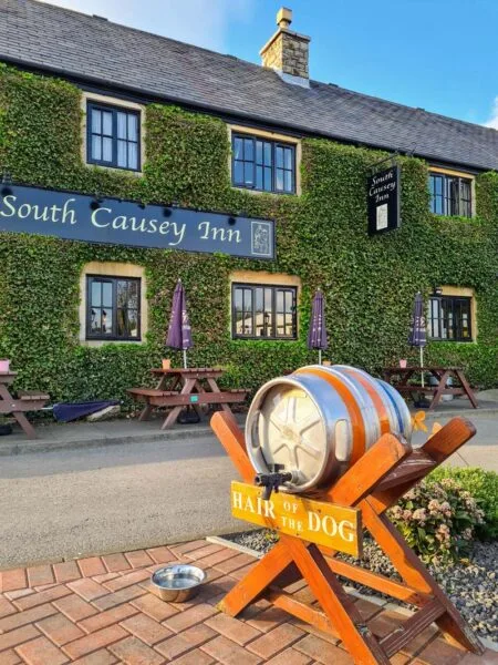 Exterior photo of South Causey Inn entrance. Ivy-covered building in background and keg placed on orange wooden stand with sign that reads "hair of the dog" and a dog bowl on the pavement. Just one of many things to do in Durham for couples