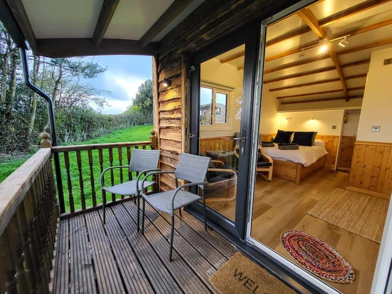 Exterior image showing deck area with two chairs in front of large glass windows, the interior of the Shepherd's Hut is visible.