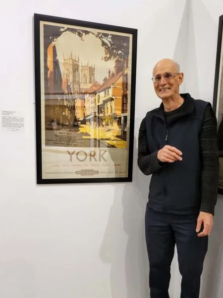 Interior photo of Terence Buckle standing next to one of his father's, Claude Buckle, railway posters. 